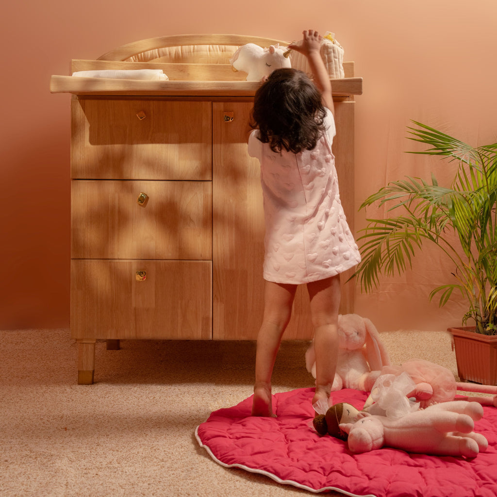 Child playing with dolls on a pink rug in front of a wooden changing table.