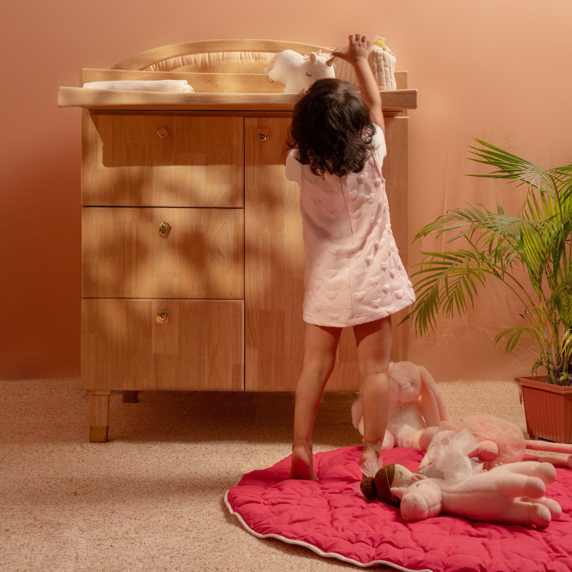 Child playing with dolls on a pink rug in front of a wooden changing table.